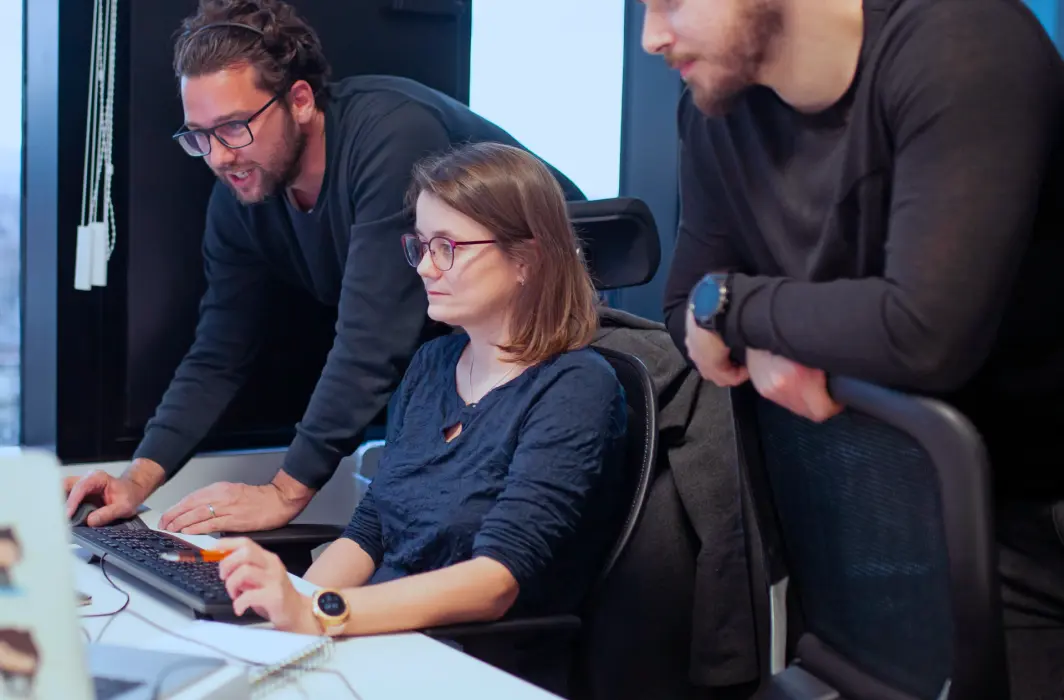 Image showing one person at a computer with two others standing beside them, pointing at something on the screen.